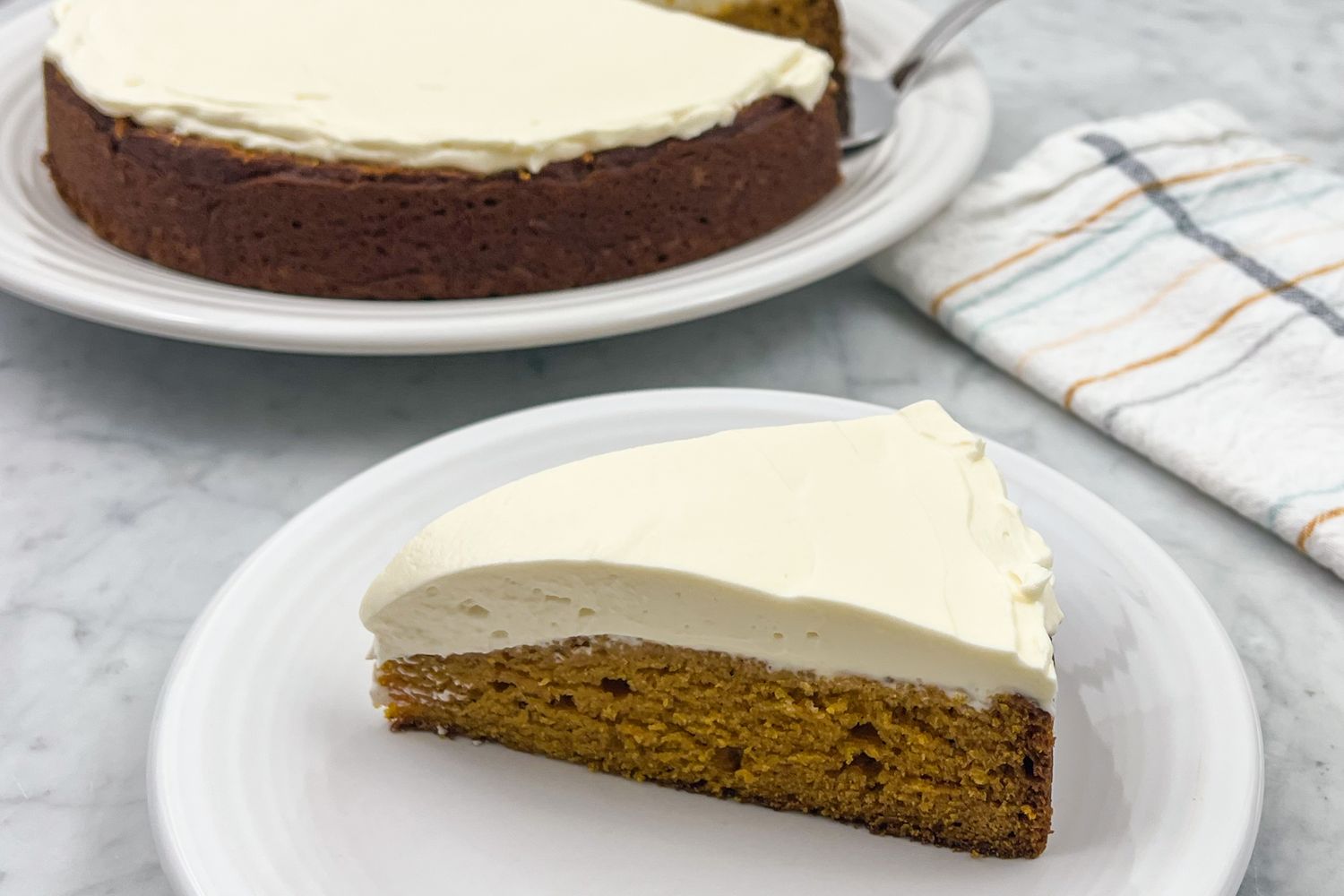 A slice of pumpkin cake with creamy topping on a white plate whole cake in the background