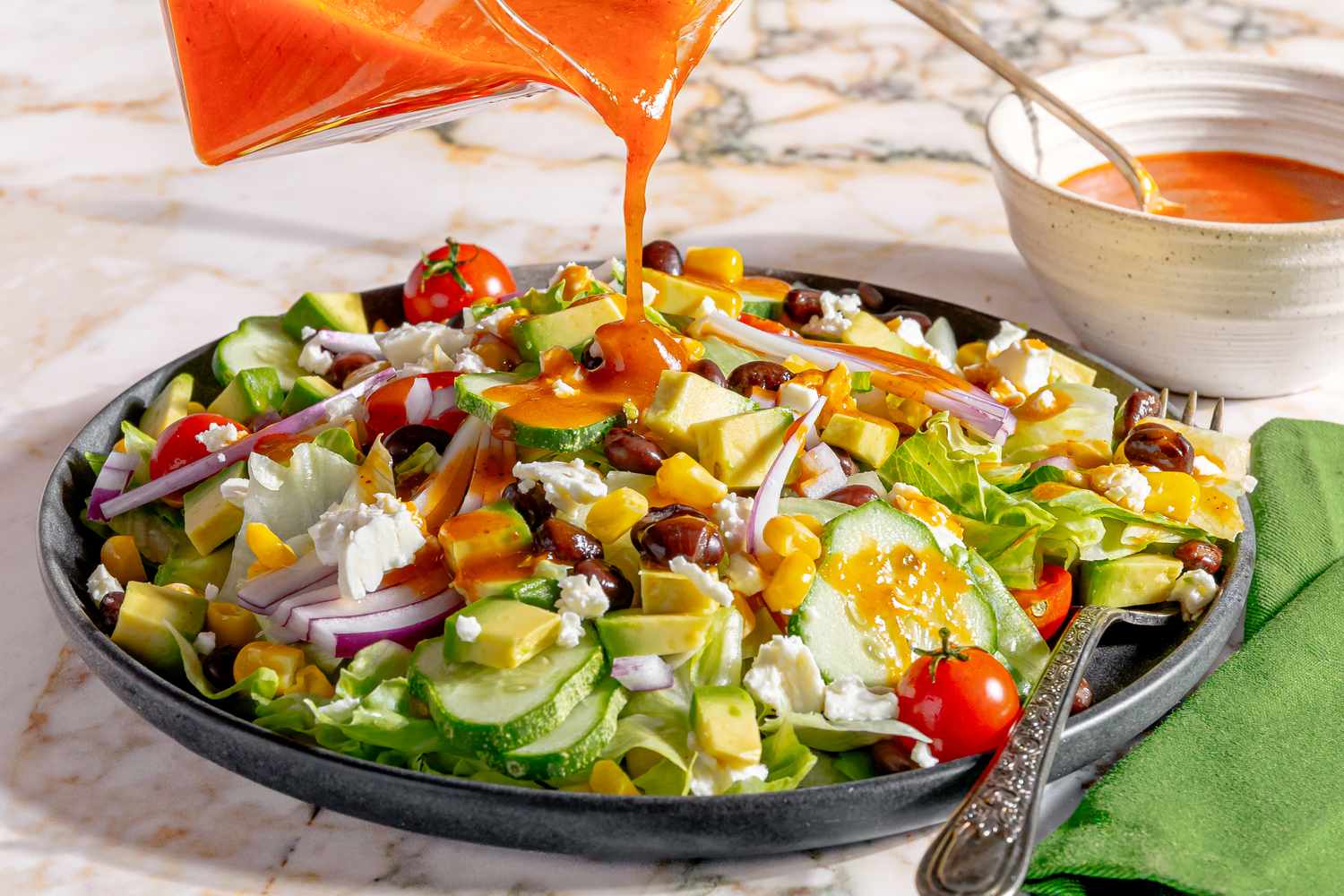 plate of salad, sauce pitcher drizzling Chipotle Honey Vinaigrette on top with a small bowl of the dressing with a spoon in the background