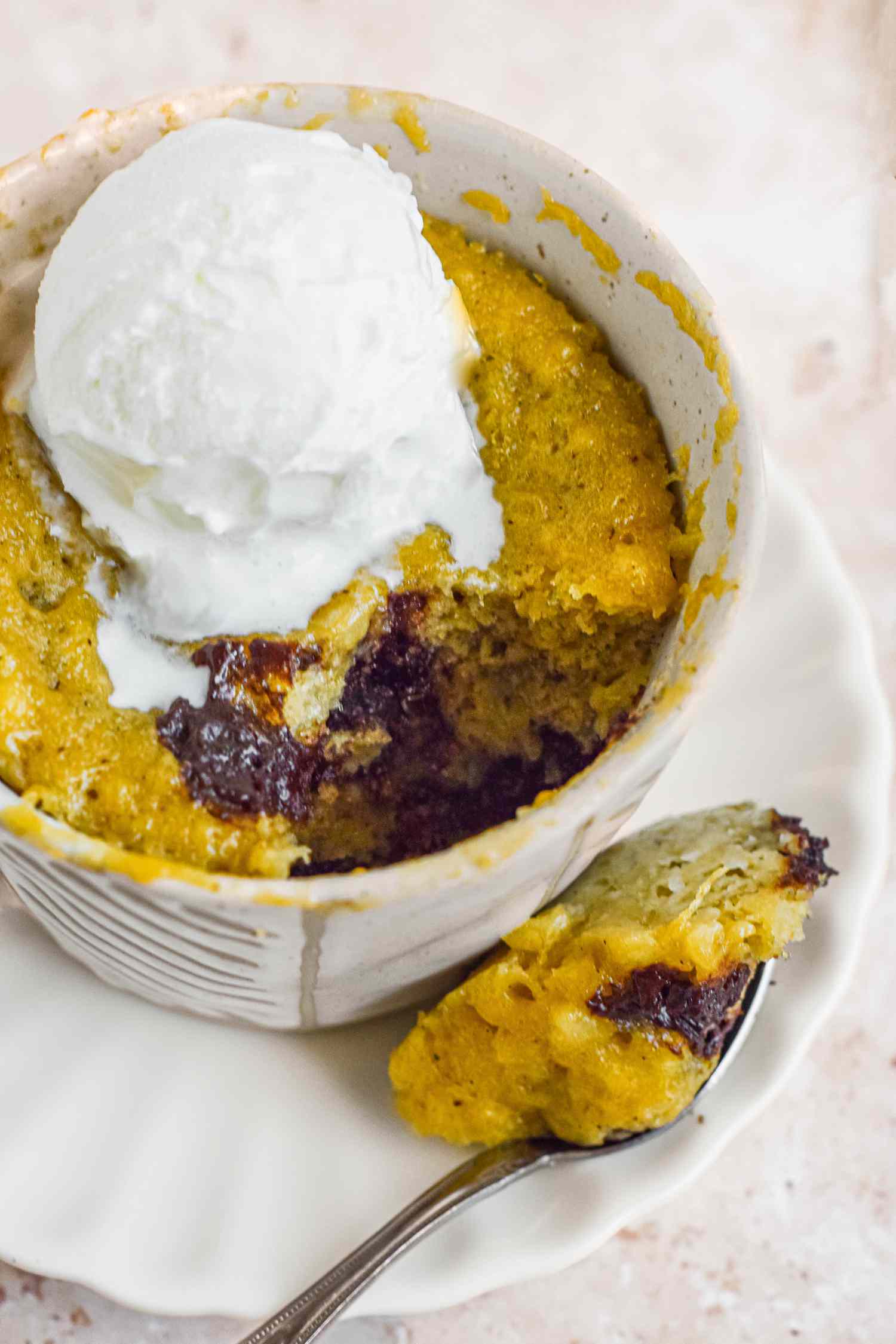 Mug of Banana Bread Chocolate Chunk Mug Cake Topped with a Dollop of Ice Cream, and on a Saucer, a Spoonful of Cake 