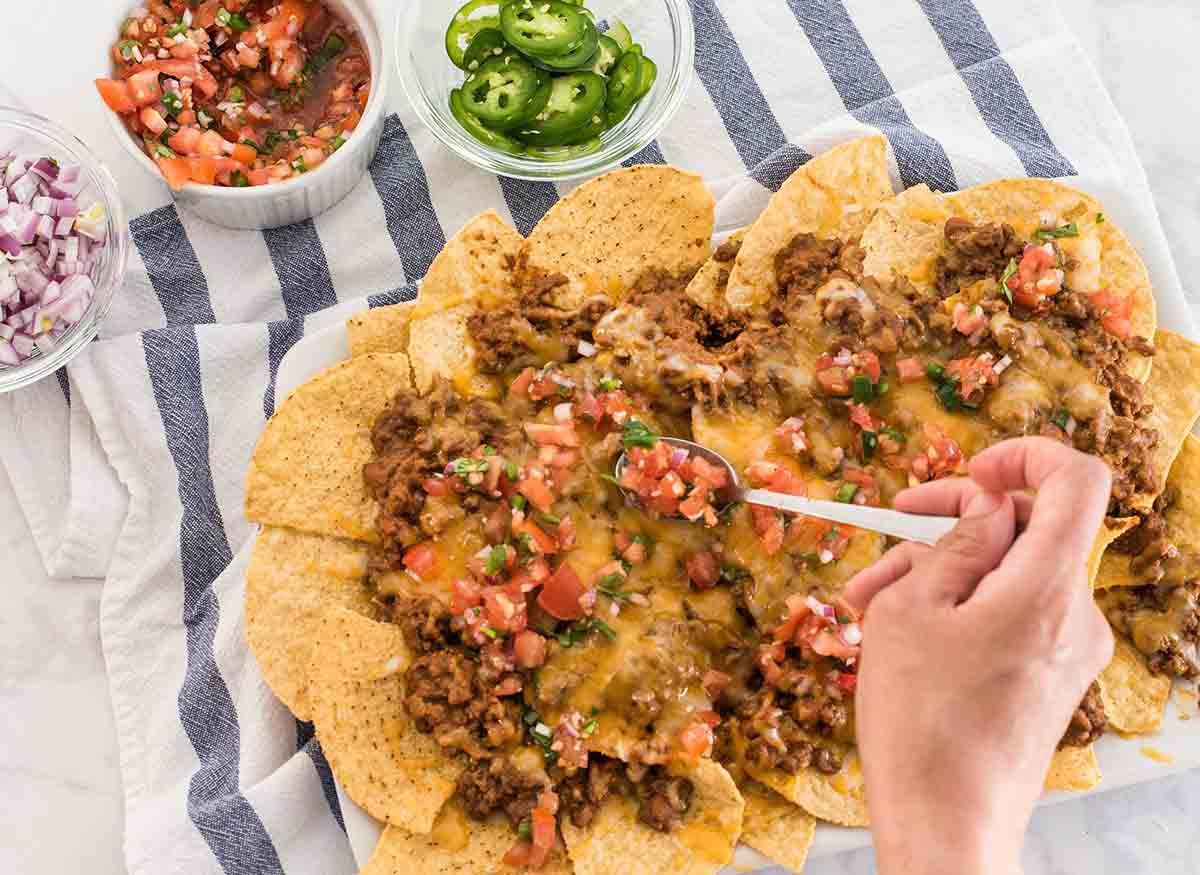 Pico de gallo being spooned onto beef and cheese nachos on a white platter 