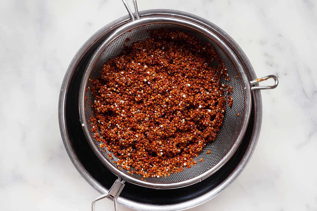 Red quinoa in a colander sitting over a metal bowl on a marble background.