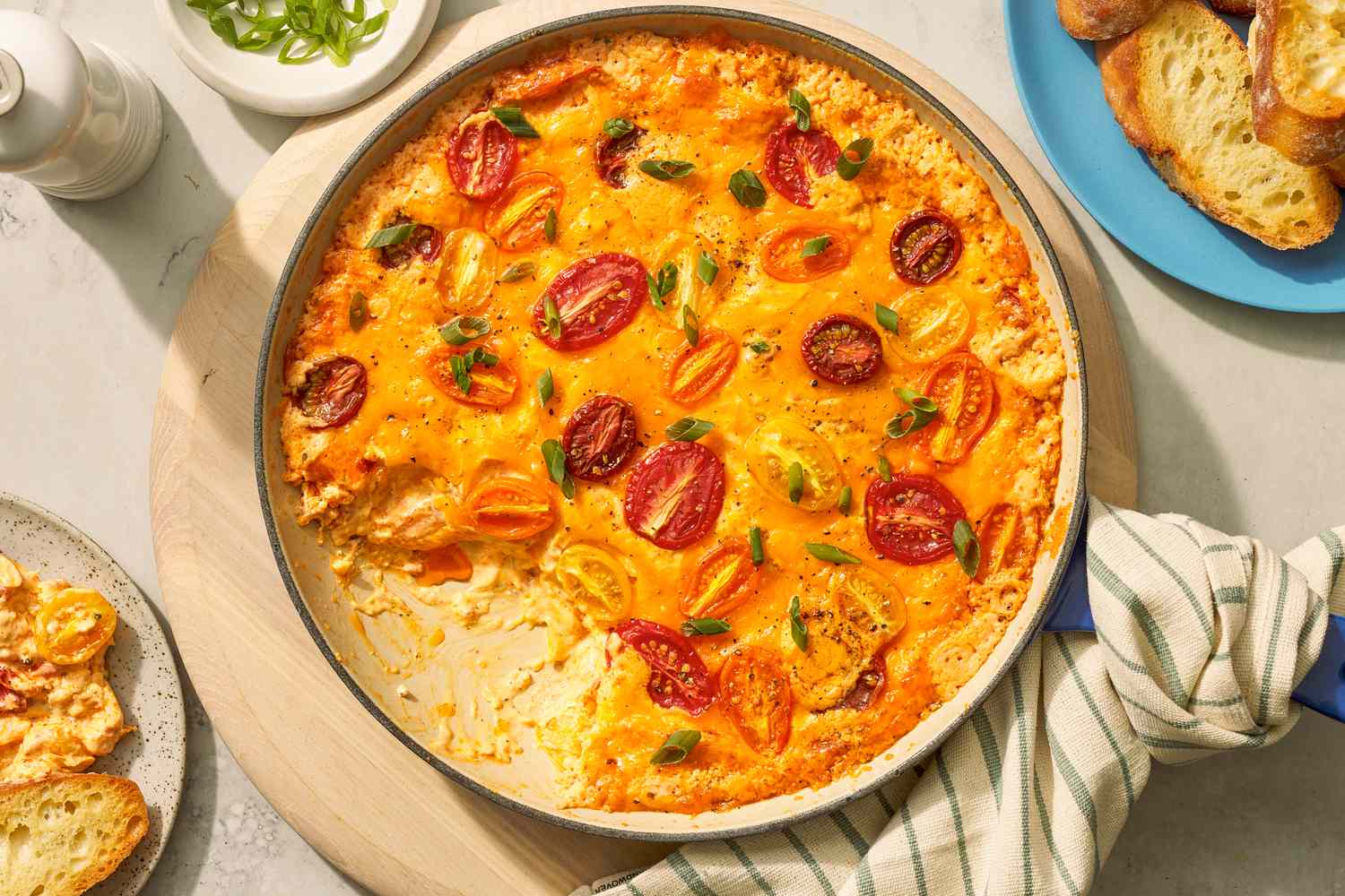 Tomato pie dip in a pan at a table setting with servings of dip on two plates with toasted baguette pieces, a bowl with sliced scallions, and a pepper grinder 
