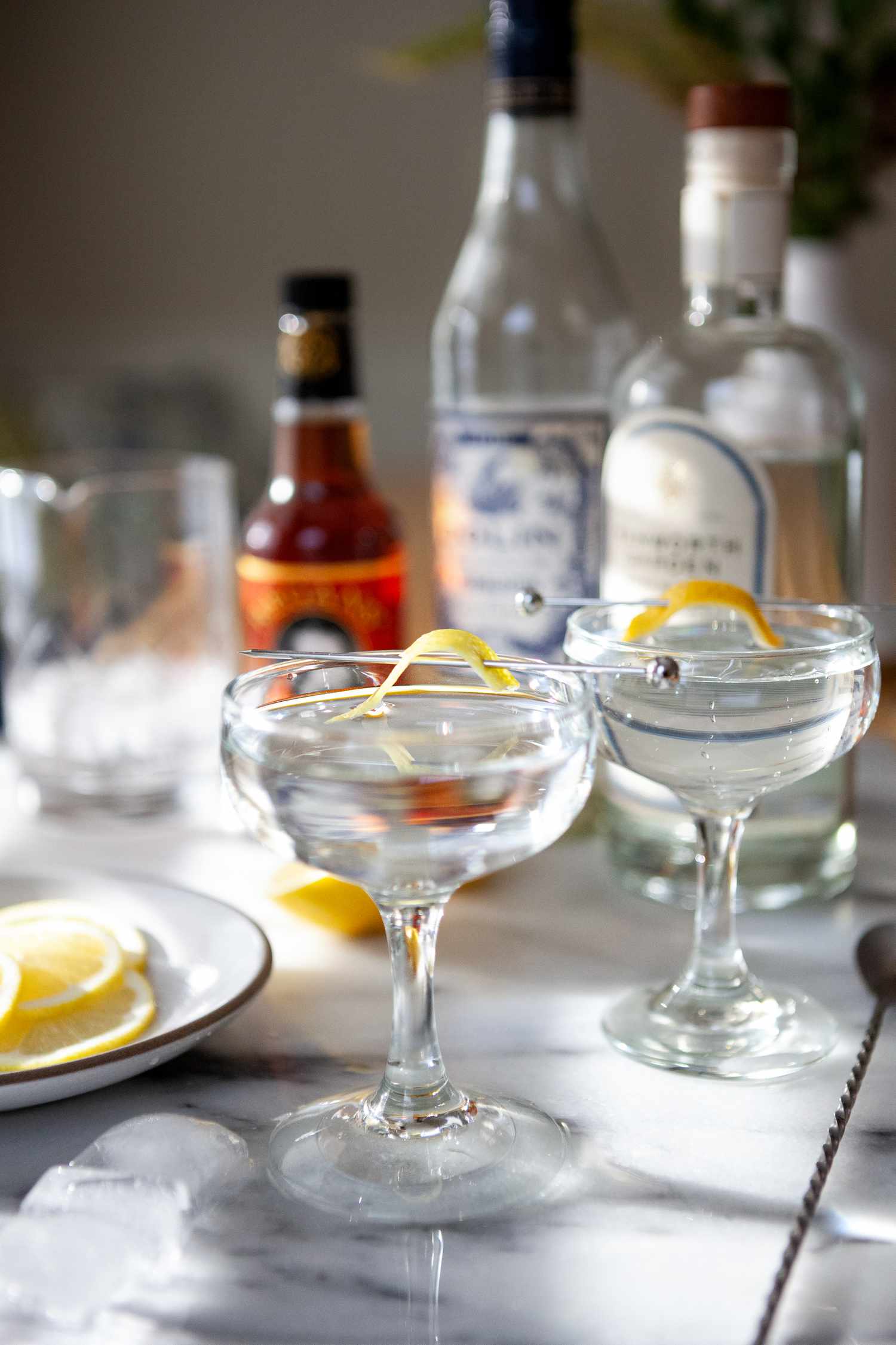 Two Classic Gin Martinis on a table with bottles and plates behind it.