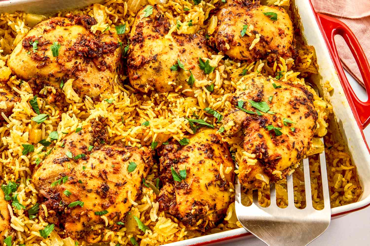Baked chicken thighs on seasoned rice in a rectangular baking dish with a serving spatula