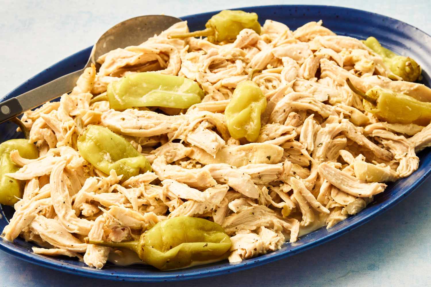 Angled view of a blue platter and a serving spoon with shredded chicken and pepperoncini