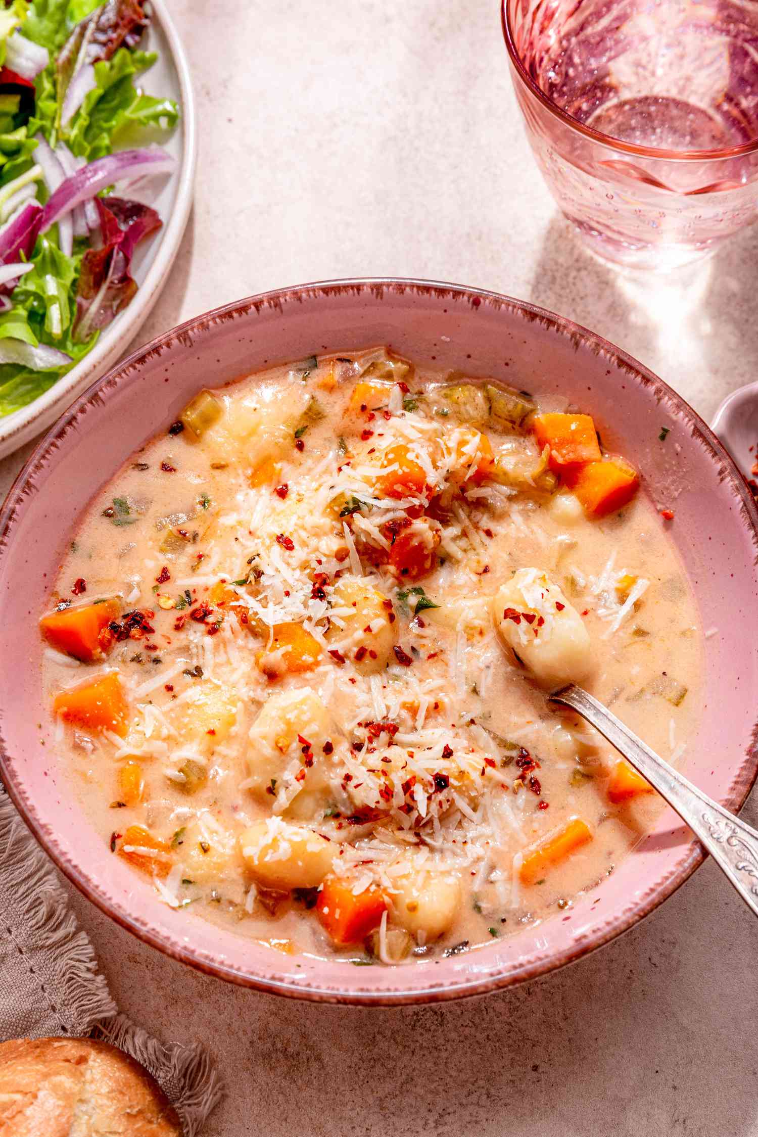 Bowl of creamy vegetable dumpling soup with a spoon, and in the surroundings, a plate of salad, a glass of water, and dinner rolls on a grey and black kitchen towel