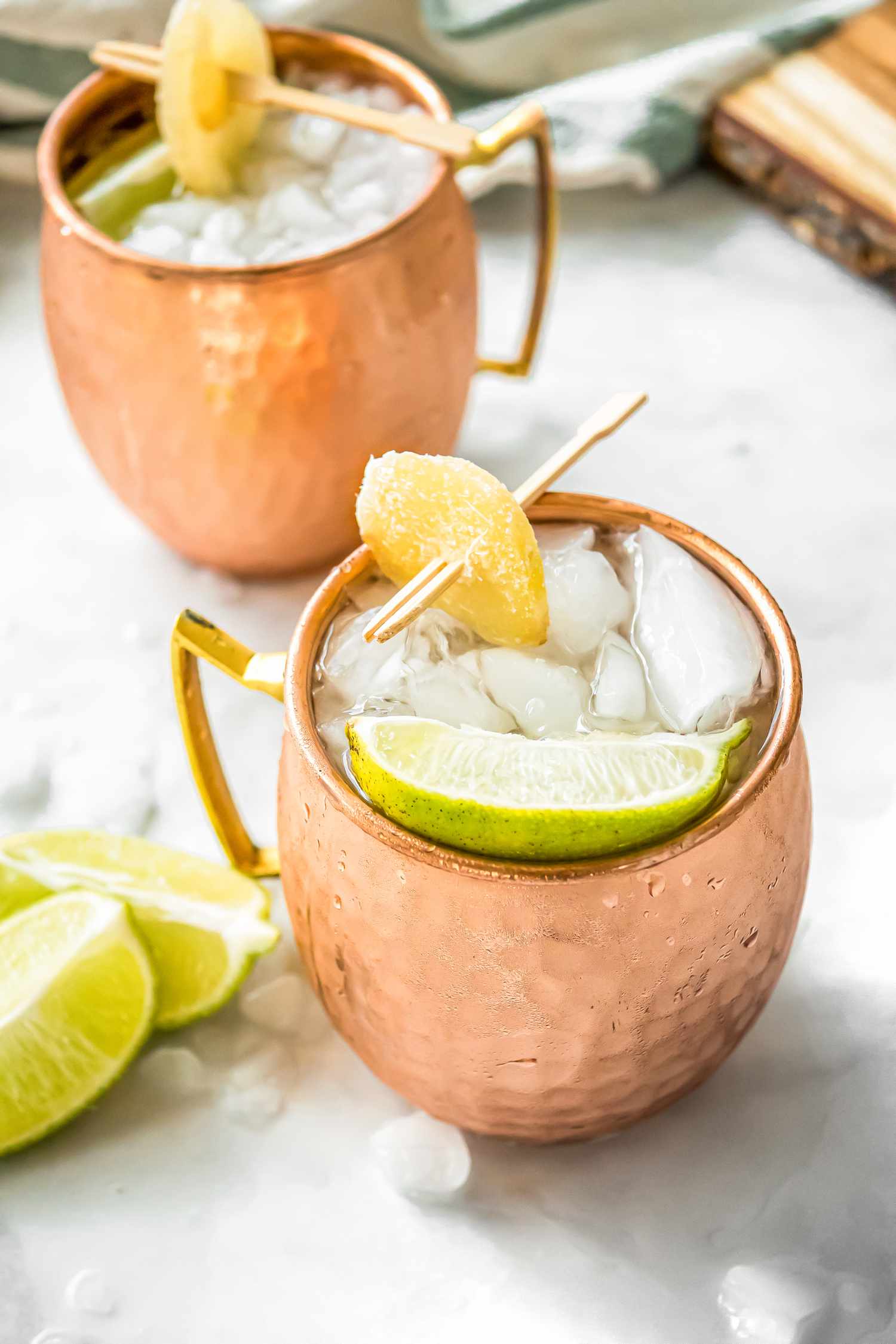 Mezcal Mule in Two Copper Mugs Garnished with Lime and Candied Ginger, Surrounded by Lime and Ice