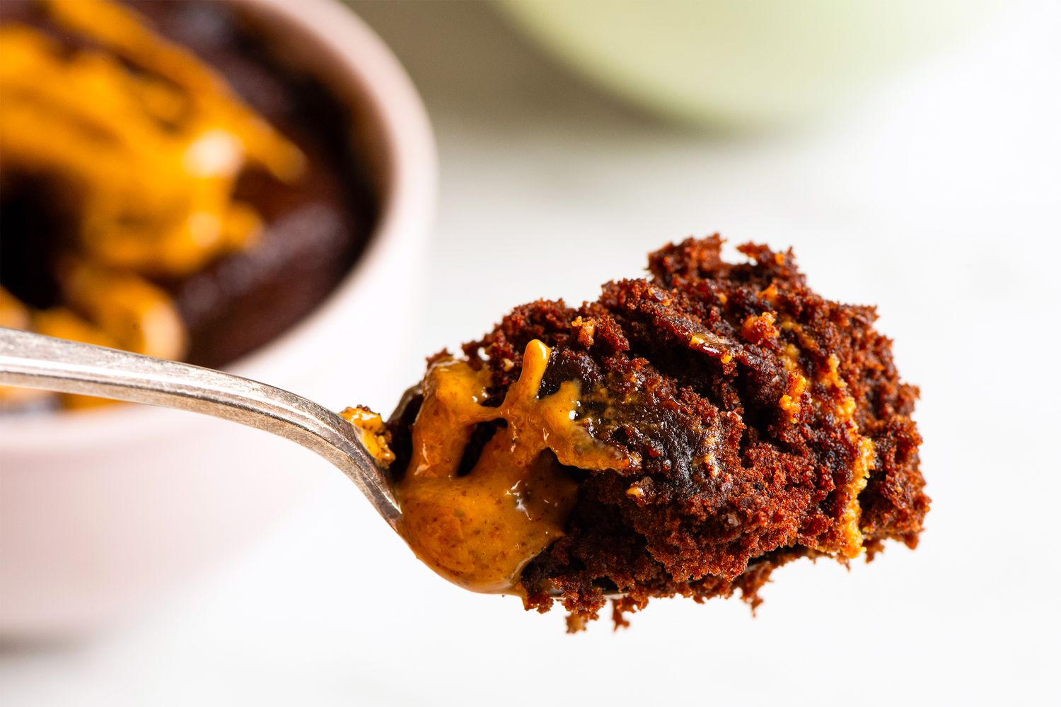 A spoon with a bite of cake topped with peanut butter in front of a bowl