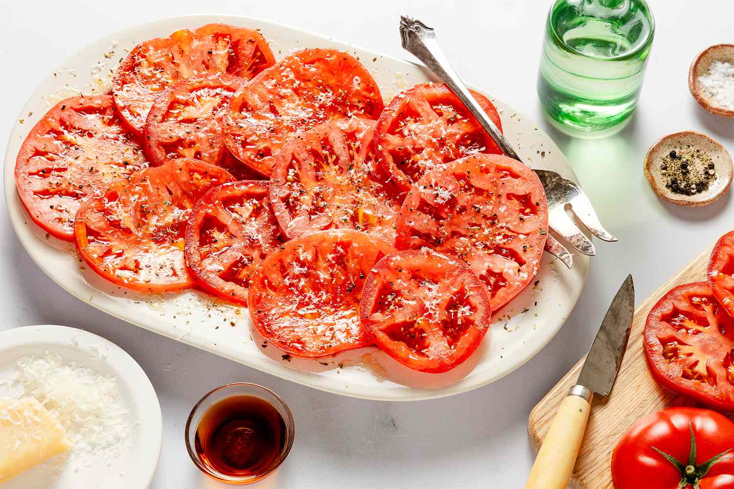 Sliced tomatoes topped with seasoning and parmesan on a white platter surrounded by garnishes and utensils on a table
