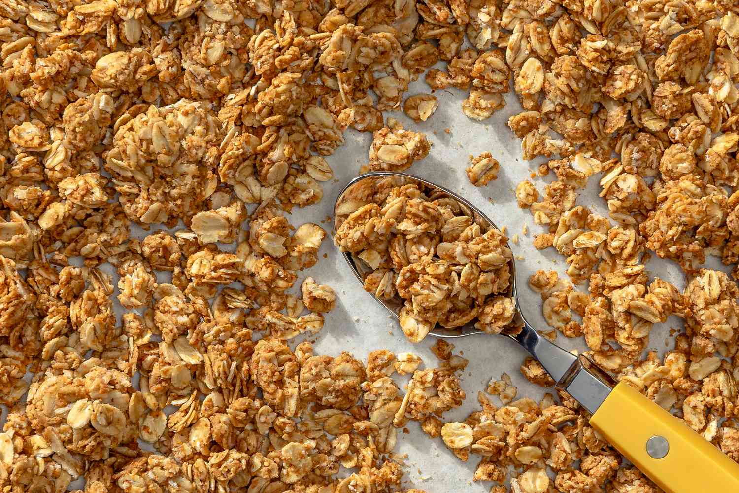 Overhead view of a metal spoon with a yellow handle in the middle of granola spread out on a light gray surface