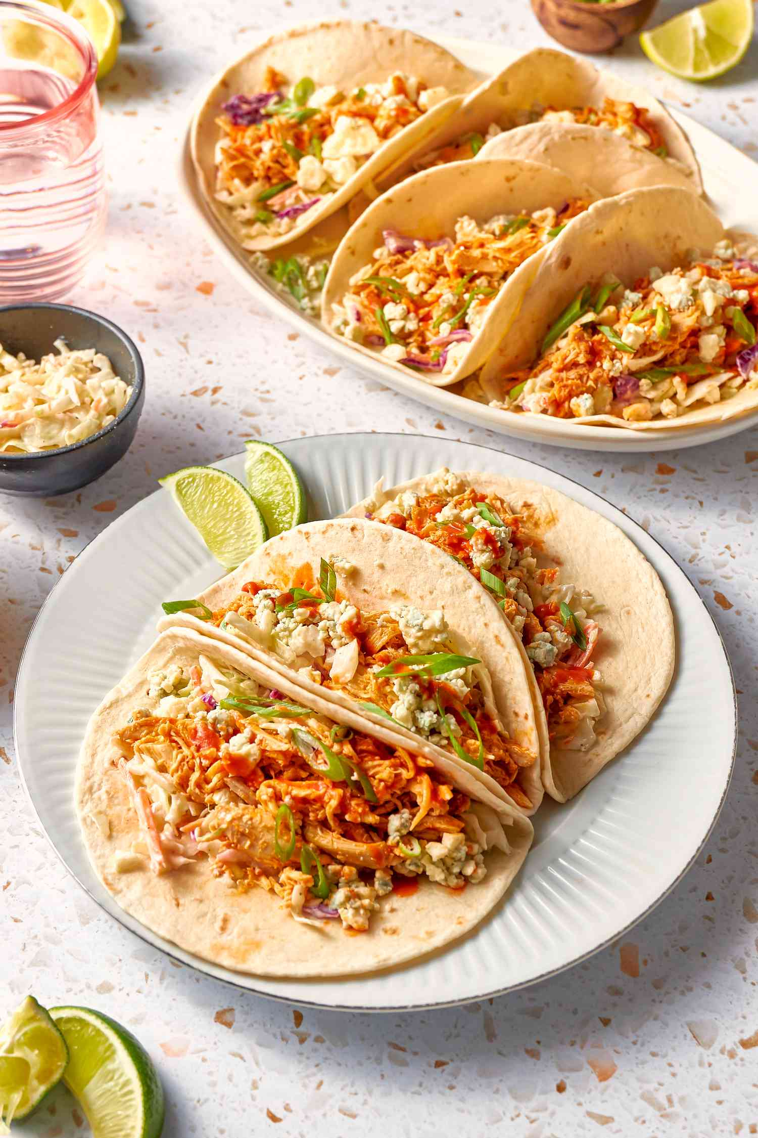 Plate With a Serving of Buffalo Chicken Tacos Topped With Blue Cheese Crumbles and Green Onion Slices and Next to Two Lime Slices. In the Surroundings, a Platter With More Tacos, Lime Slices on the Counter, a Bowl of Coleslaw, and a Glass 