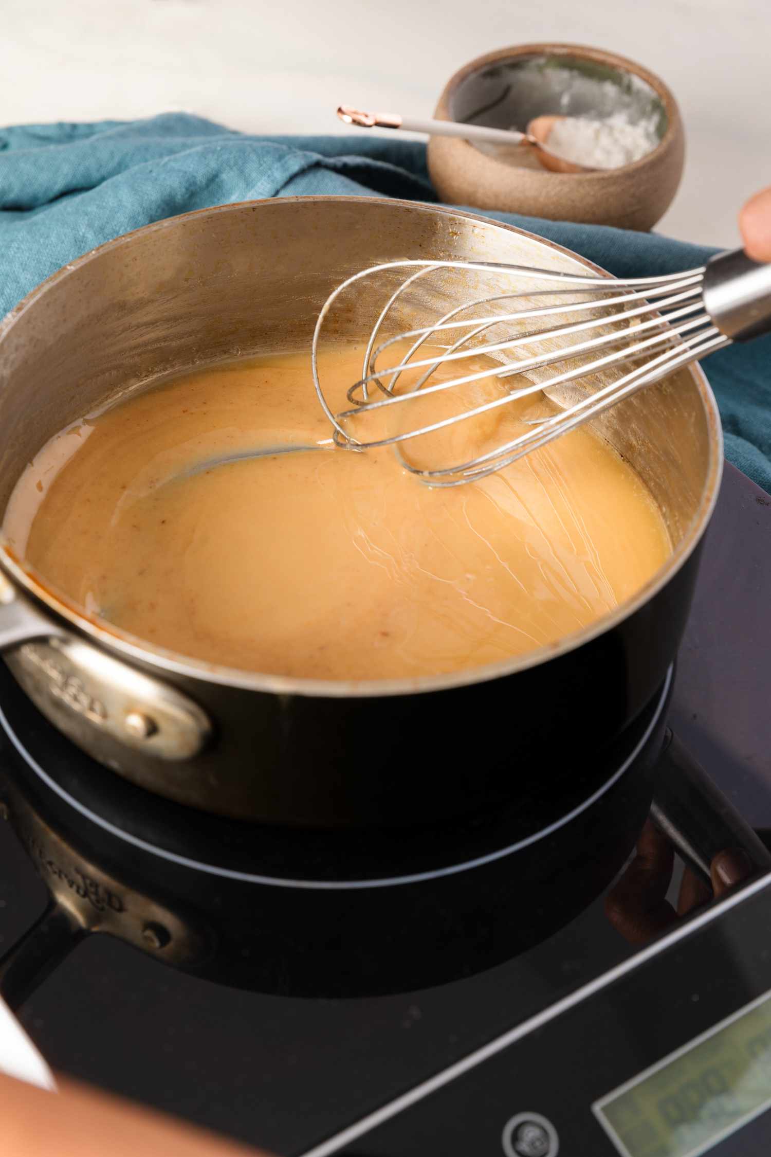 Roux whisked in a saucepan on an induction stove, and in the surroundings, a bowl of flour and a blue kitchen towel