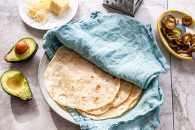 Homemade flour tortillas on a plate in-between a teal linen. A small bowl of pickled jalepanos, a box shredder, shredded sheese and a halved avocado are around the plate.