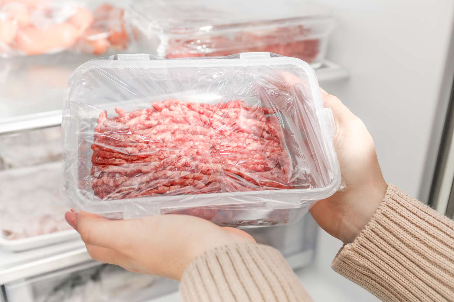 Hands holding a plastic container with ground meat wrapped in plastic, with a refrigerator shelf in the background
