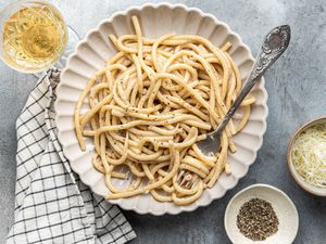 Plate of Brown Butter Cacio e Pepe with Some Noodles Wrapped around a Fork, and Next to It, a Kitchen Towel, a Glass of Wine, and Two Small Bowls (One with Cracked Black Pepper and Another with Shredded Parmesan) 