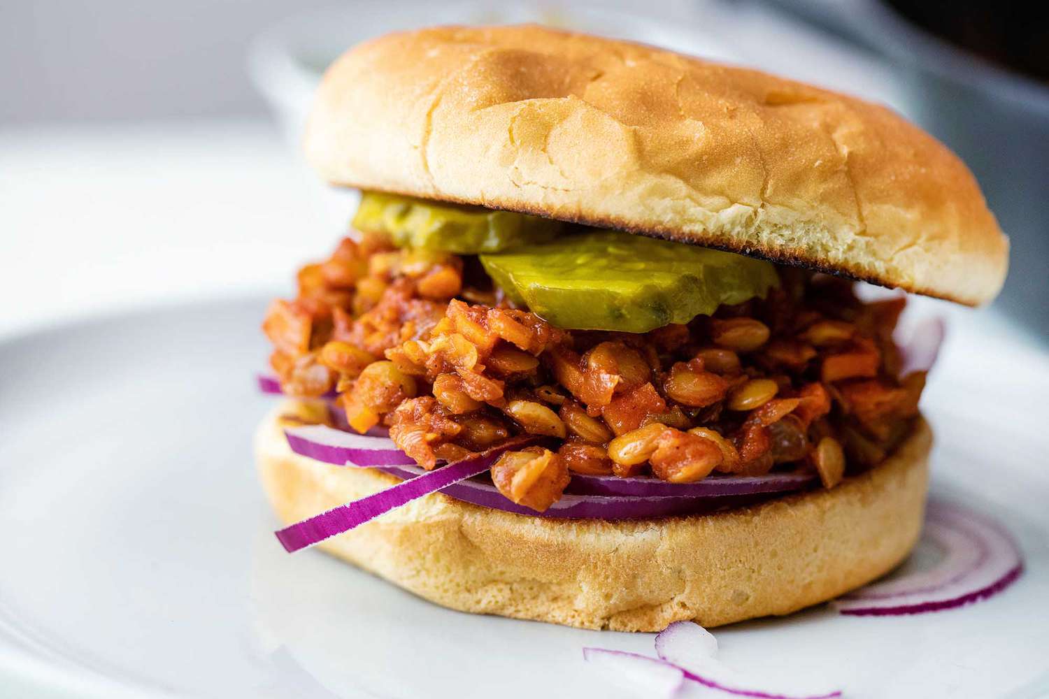 A lentil sloppy joe sandwich on a plate with red onion and pickle slices.