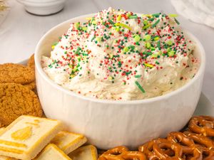 A bowl of festive dessert dip topped with sprinkles surrounded by cookies and pretzels on a serving tray