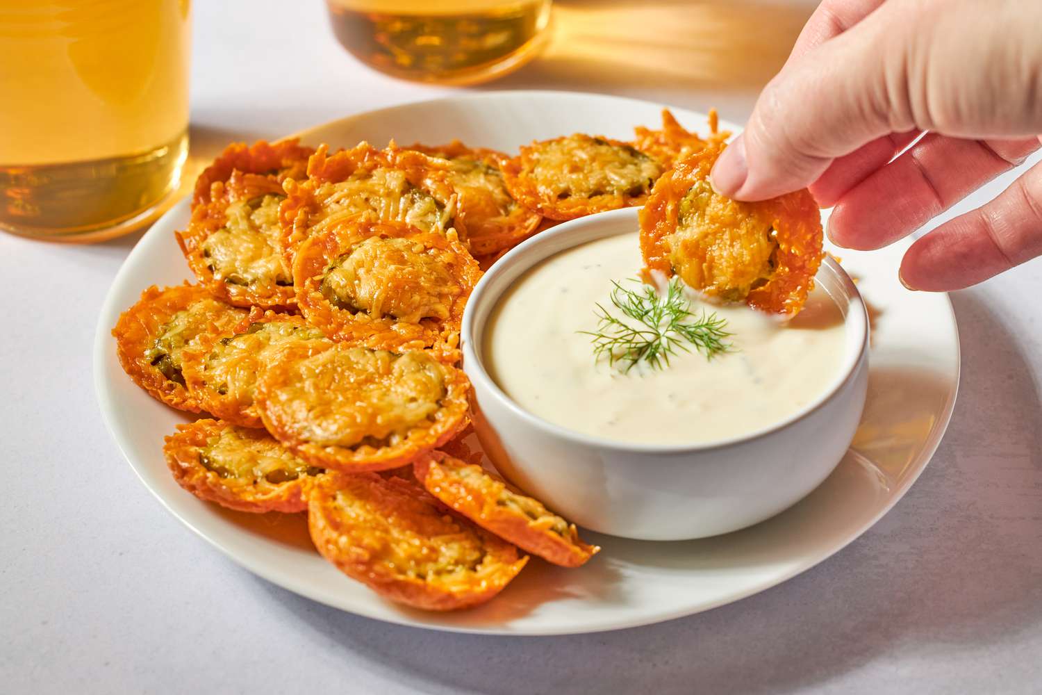 A plate of crispy fried pickles with a hand dipping one into a bowl of white sauce, garnished with a sprig of dill