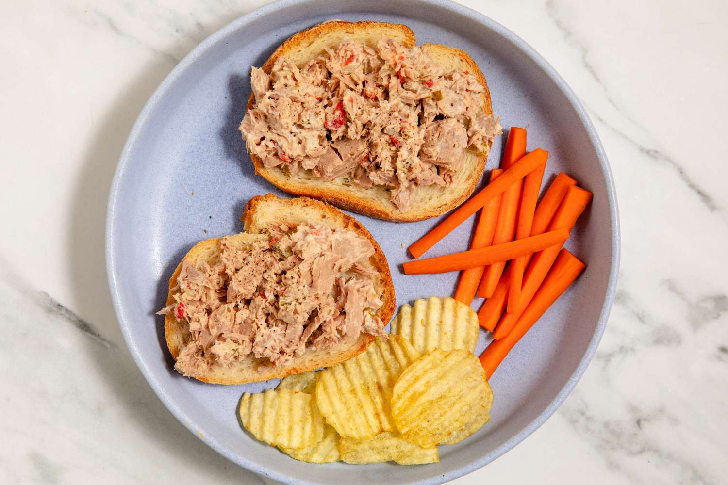 Two slices of bread with tuna salad, next to carrot sticks and potato chips on a plate