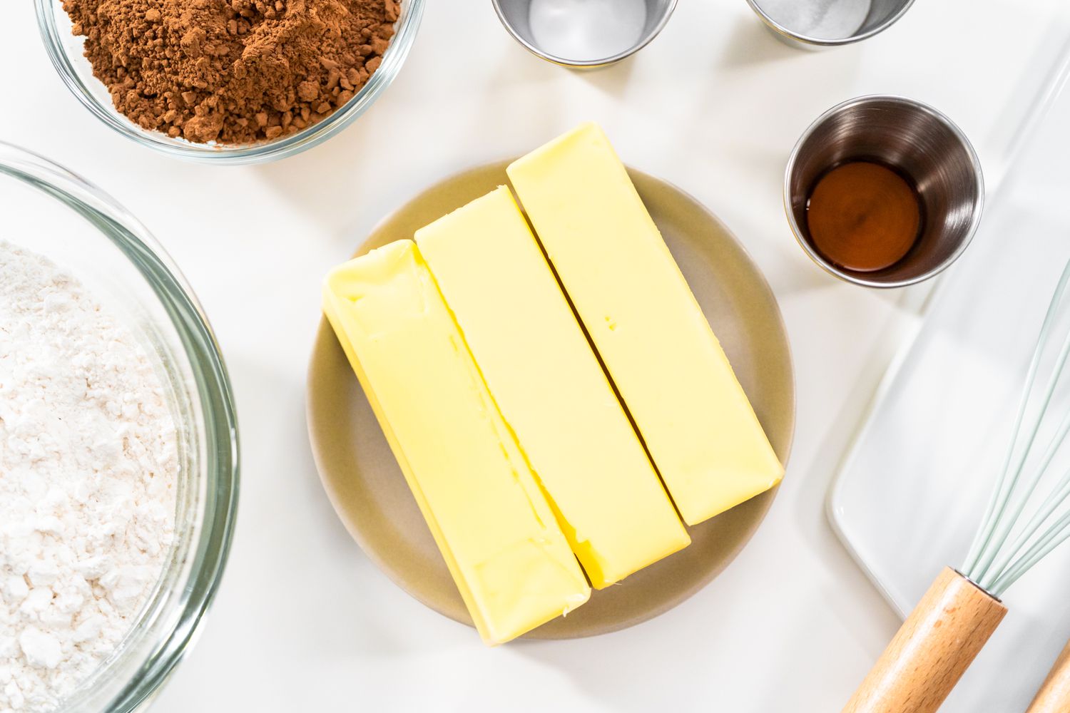 A baking scene showing ingredients laid out on a countertop—sticks of butter, vanilla, cocoa powder, and flour