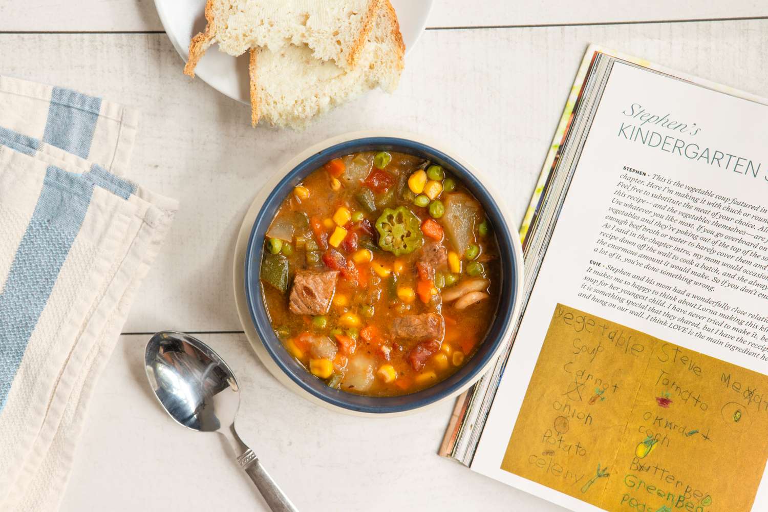 A bowl of vegetable beef soup next to a magazine or cookbook and slices of bread with a spoon placed on the table beside it