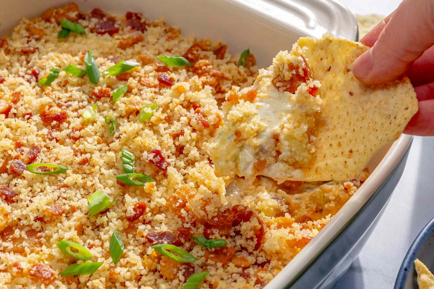 Closeup of a baking dish with jalapeño popper dip, and a hand holding a tortilla chip scooping some of the dip out
