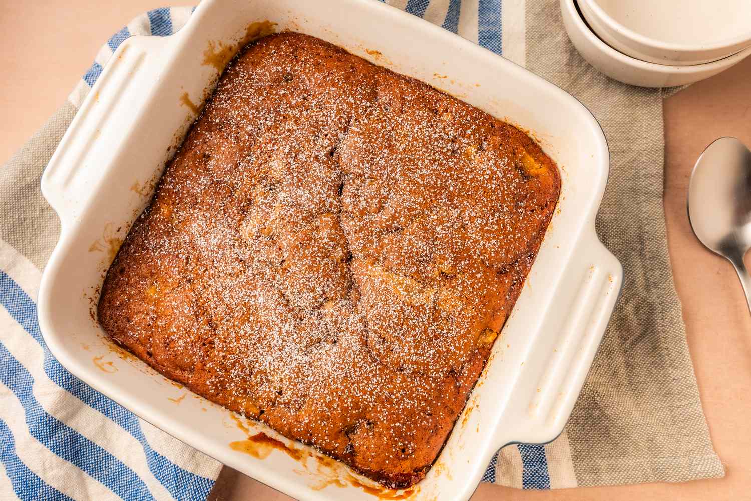 Square baking dish with a pudding cake lightly dusted with powdered sugar on a striped cloth beside a spoon and stacked plates