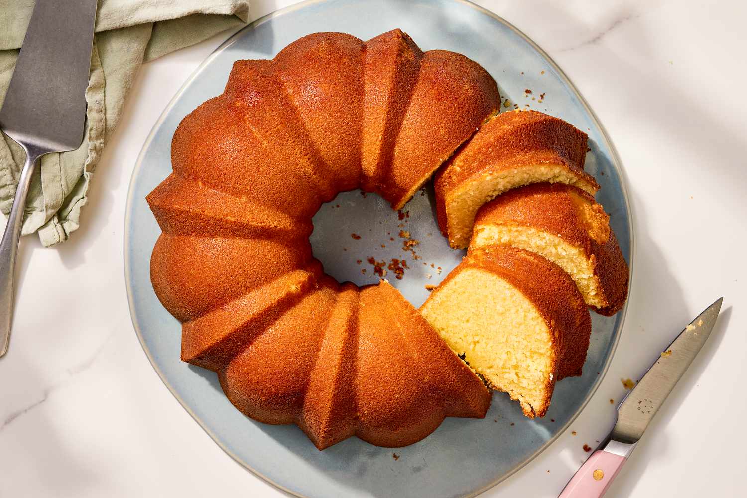 Overhead view of a light blue plate with a partially sliced pound cake next to a cake server and knife on a marble countertop