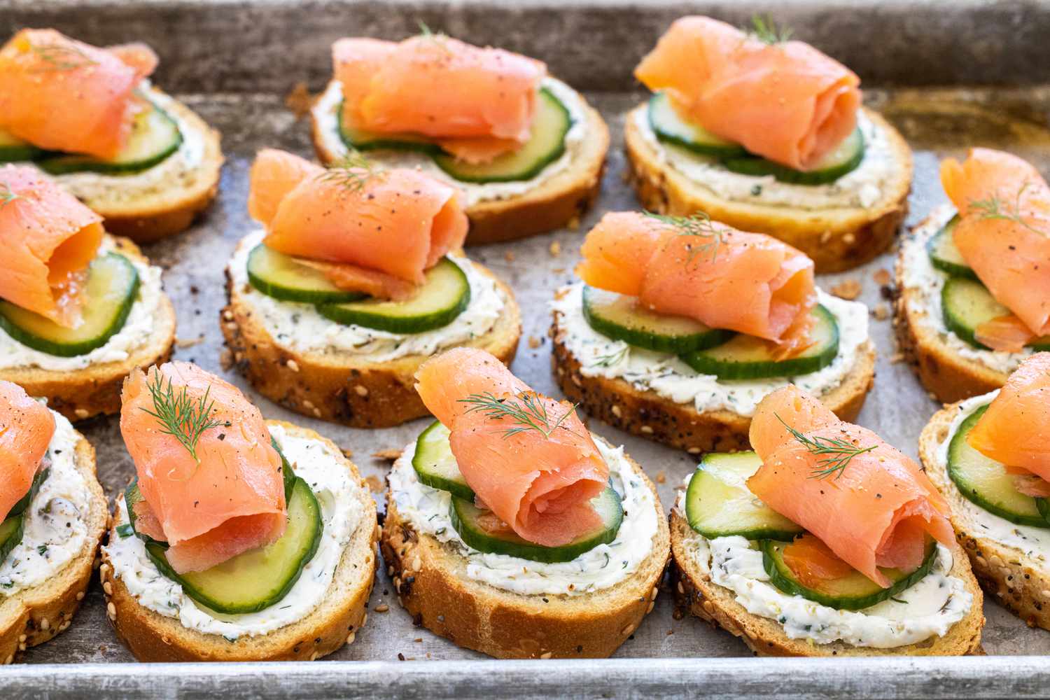 Crostini topped with smoke salmon, pickled cucumbers, and dilled cream cheese on a baking sheet.