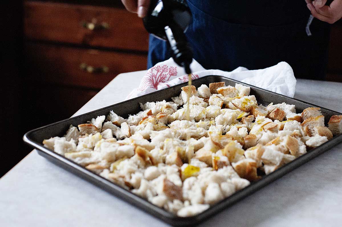 Woman drizzling olive oil on cubed bread on a baking sheet to make thanksgiving stuffing wiht apples and sausage..