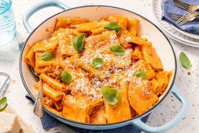 Pot of Paccheri With Quick Sausage Ragu With a Spoon, and in the Surroundings, a Glass, a Block of Parmesan, a Stack of Plates, and Basil Leaves