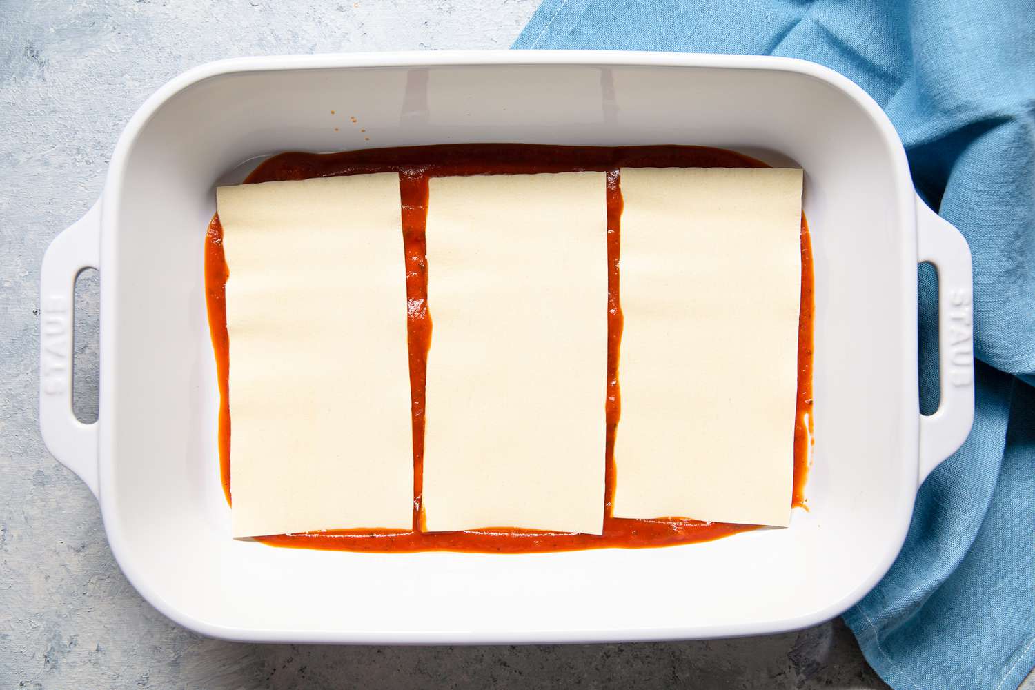 Oven-Ready Lasagna Noodles Added to First Layer of Marinara Sauce in the Casserole Dish Next to the Sky Blue Kitchen Towel