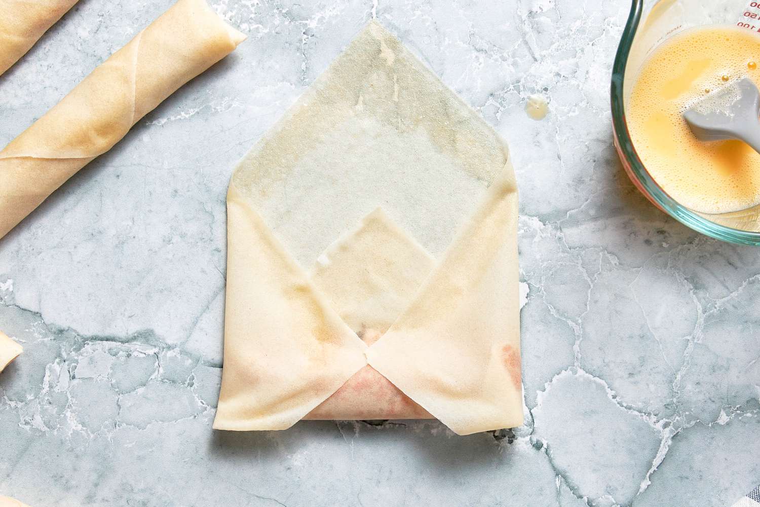 Left and Right Edges of Egg Roll Wrapper Folded Over the Filling (Making an Envelope Shape), and in the Surroundings, and in the Surroundings, Rolled Lumpia and a Pyrex Measuring Cup With the Egg Wash