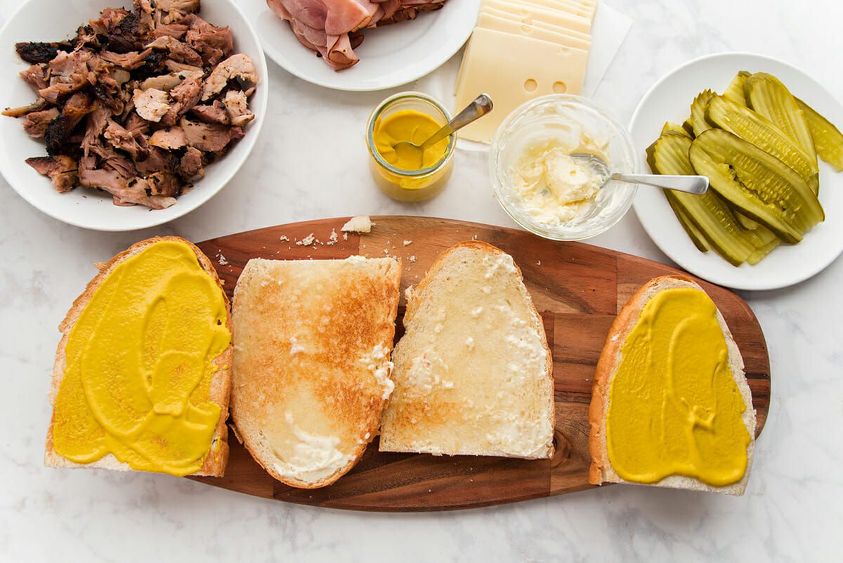 Toasted french bread is cut into quarters and set on a wooden cutting board. The middle pieces are spread with mayonnaise and the outer quarters are spread with yellow mustard. Bowls of shredded pork, sliced ham, yellow mustard, sliced cheese, mayonnaise and sliced pickles are set above.