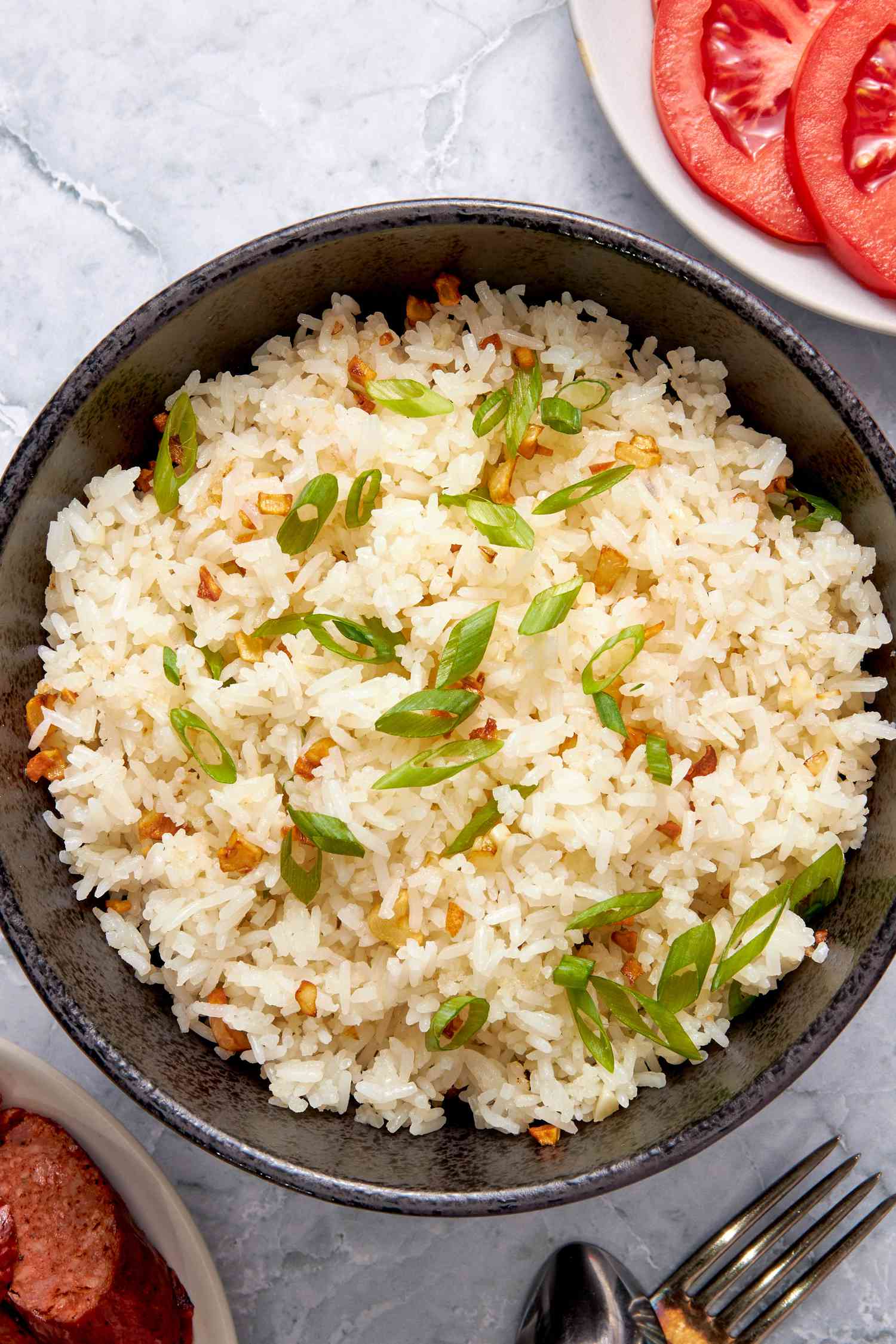 Bowl of Garlic Fried Rice (Sinangag na Kanin) next to some utensils, a plate of sliced longanisa (filipino sausage) and sliced tomatoes