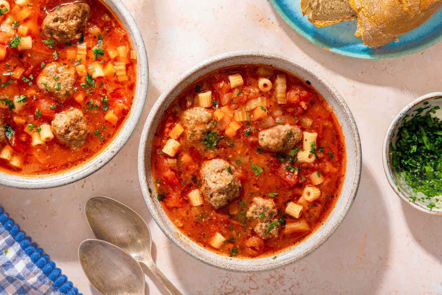 Overhead view of a two bowls of Italian meatball soup next to two spoons, a plate of bread and small bowl of parsley
