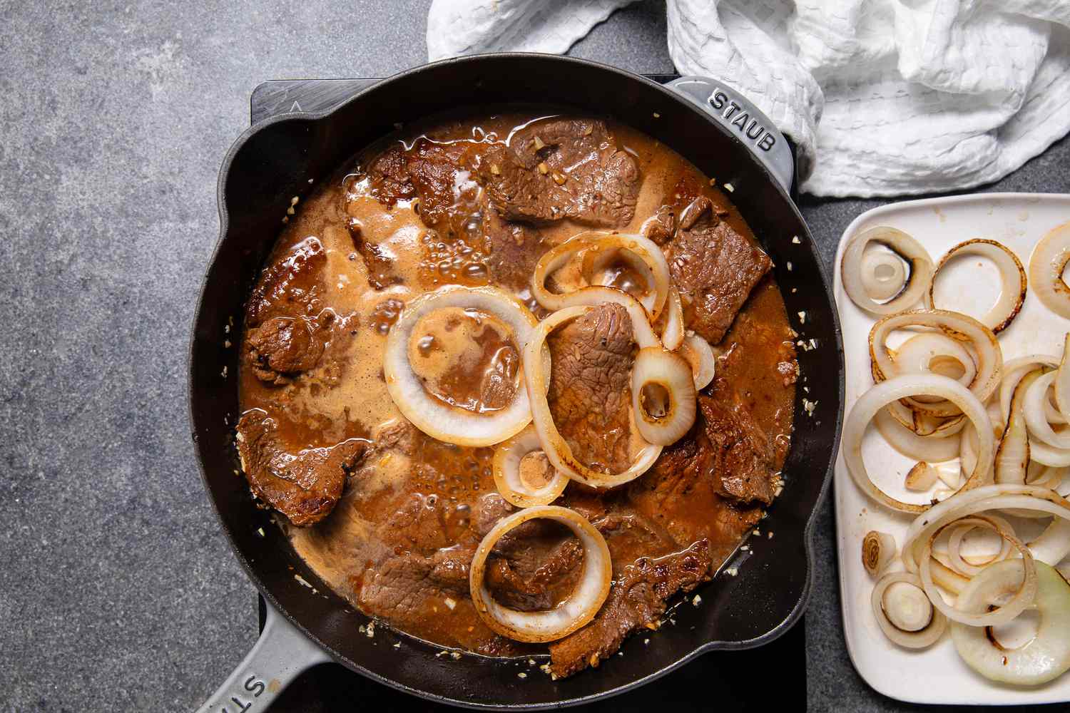 Bistek and Onions Cooking in the Marinade in the Cast Iron Skillet Next to the Plate With Onions and a White Kitchen Towel