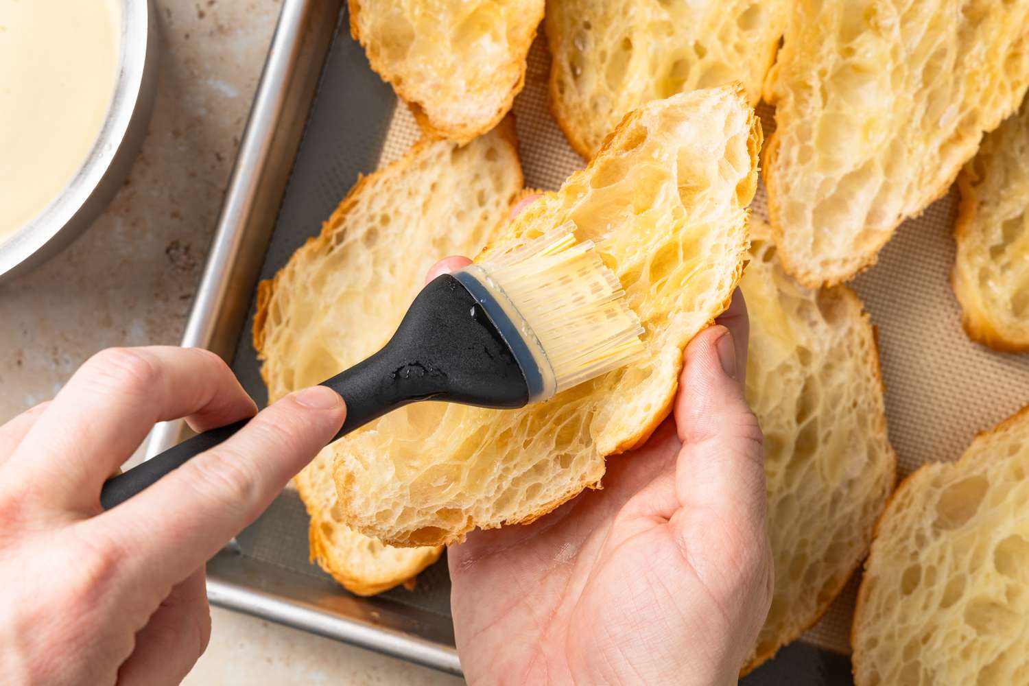 Simple Syrup Brushed on a Croissant Half, and in the Background, the Rest of the Croissant Halves on the Baking Sheet and a Bowl of Simple Syrup
