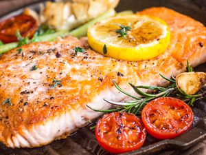Close-up of a cooked salmon fillet in a grill pan with a lemon slice on top and a halved cherry tomato and rosemary sprig next to it