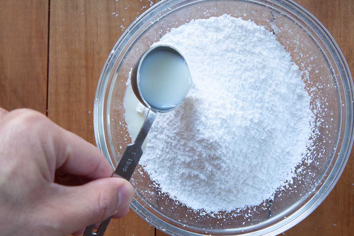 A tablespoon of milk is being added to the bowl of sifted powdered sugar.