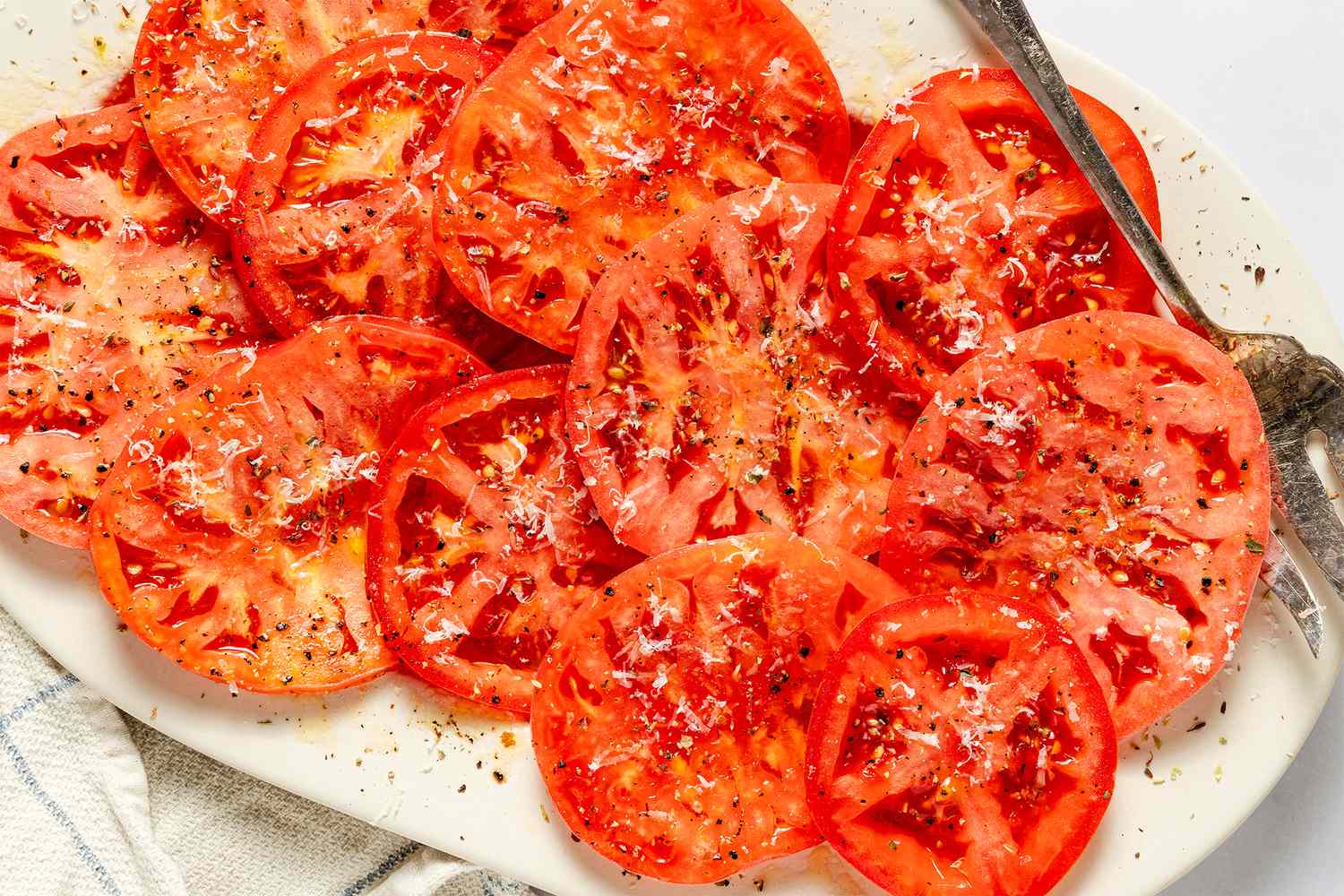 Plated marinated tomato slices topped with seasoning and shredded cheese served on a white dish with a fork