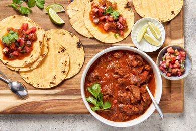 On a wooden board, a bowl of carne picada with a sprig of cilantro and a spoon, surrounded by a bowl of pico de gallo, a small plate of lime wedges, and tortillas spread all over the board (some with carne picada and pico de gallo, some plain)