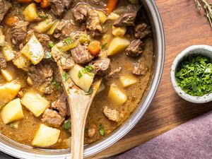A pot of beef stew with carrots and potatoes a wooden spoon placed inside a bowl of herbs in the background