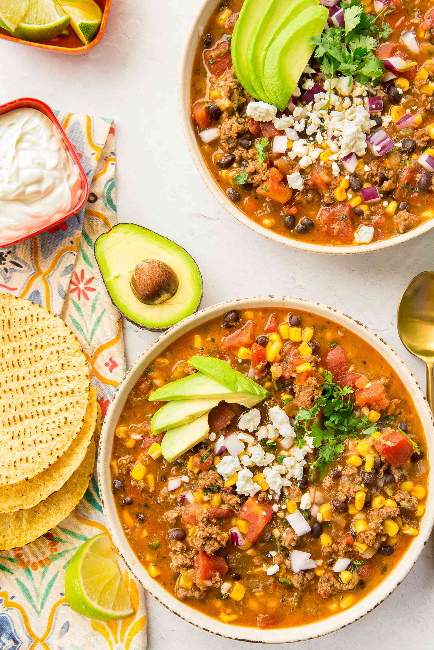 Two Bowls of Stovetop Taco Soup Topped with Avocado Slices, Cotija Cheese, and Cilantro and Surrounded by Bowls of Toppings