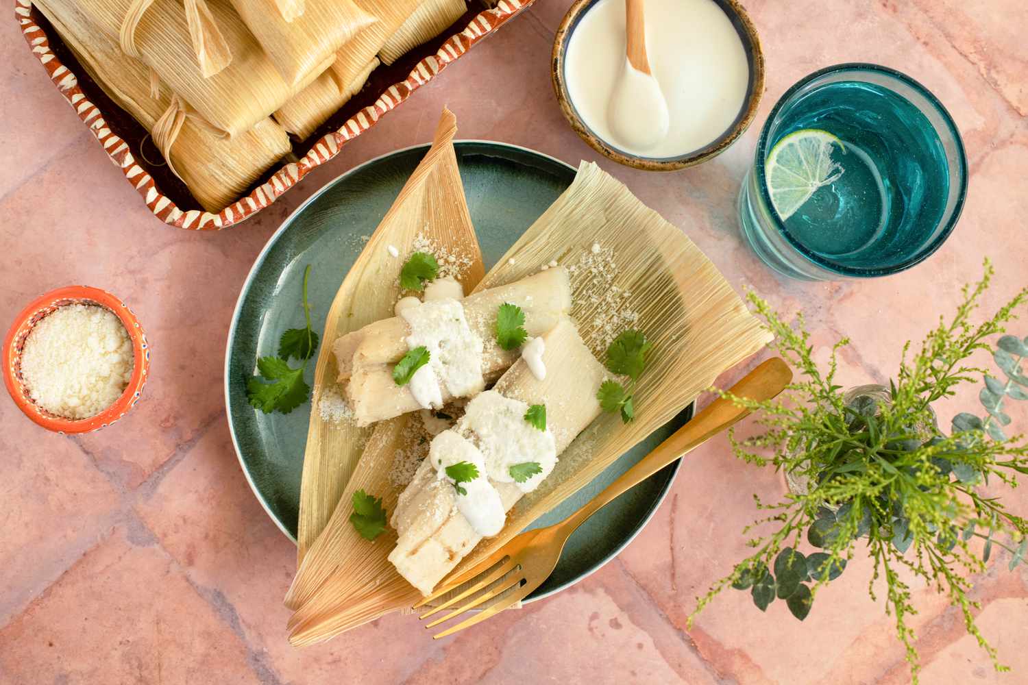 A Plate of Chicken Tamales with Chile Verde Topped with Crema, Cotija Cheese, and Some Cilantro at a Table Setting with a Platter of More Tamales, Utensils, a Small Bowl of Cotija Cheese, a Small Bowl of Crema, a Glass of Water, and a Small Vase