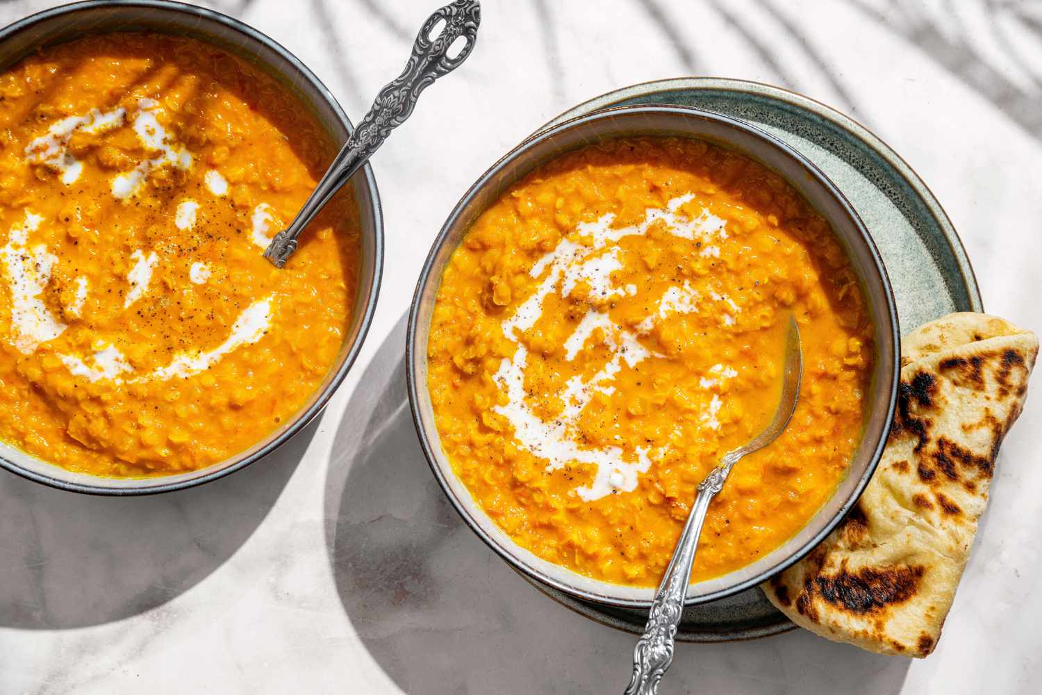 Coconut Lentil Soup in two bowls with spoons, garnished with yogurt sauce, naan on the side