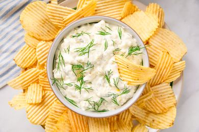 A bowl of dip garnished with dill surrounded by potato chips arranged on a plate
