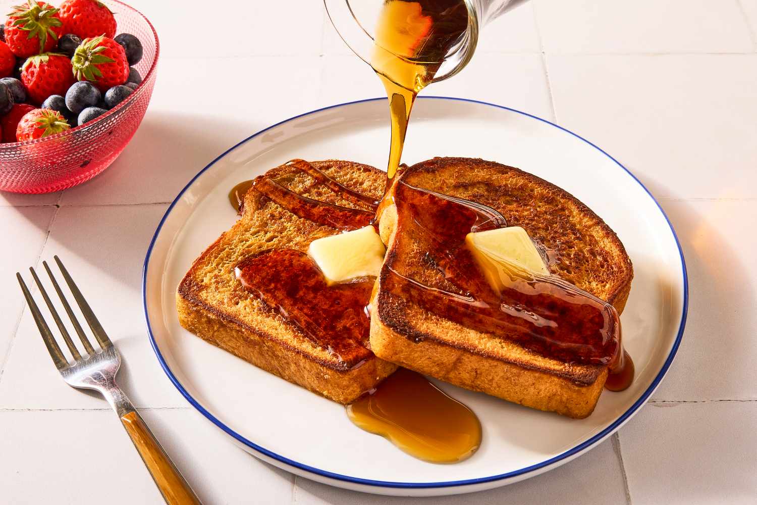 French toast slices on a plate with butter syrup being poured bowl of berries and fork nearby