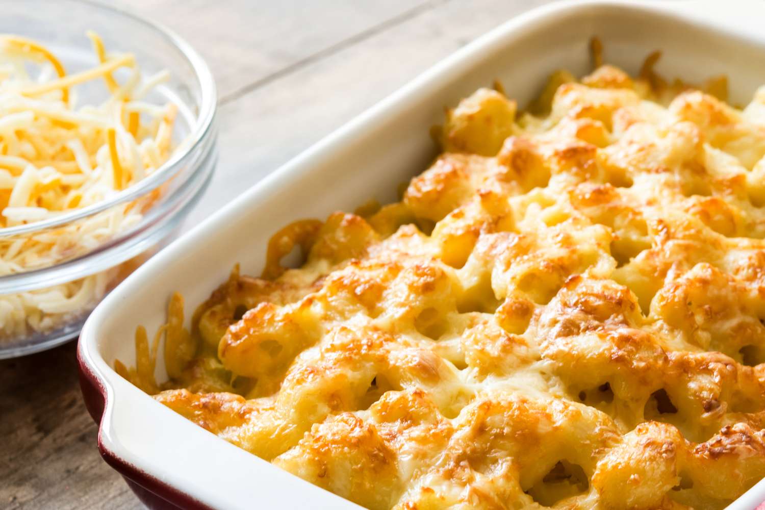 Baked macaroni and cheese in a casserole dish with a bowl of shredded cheese beside it