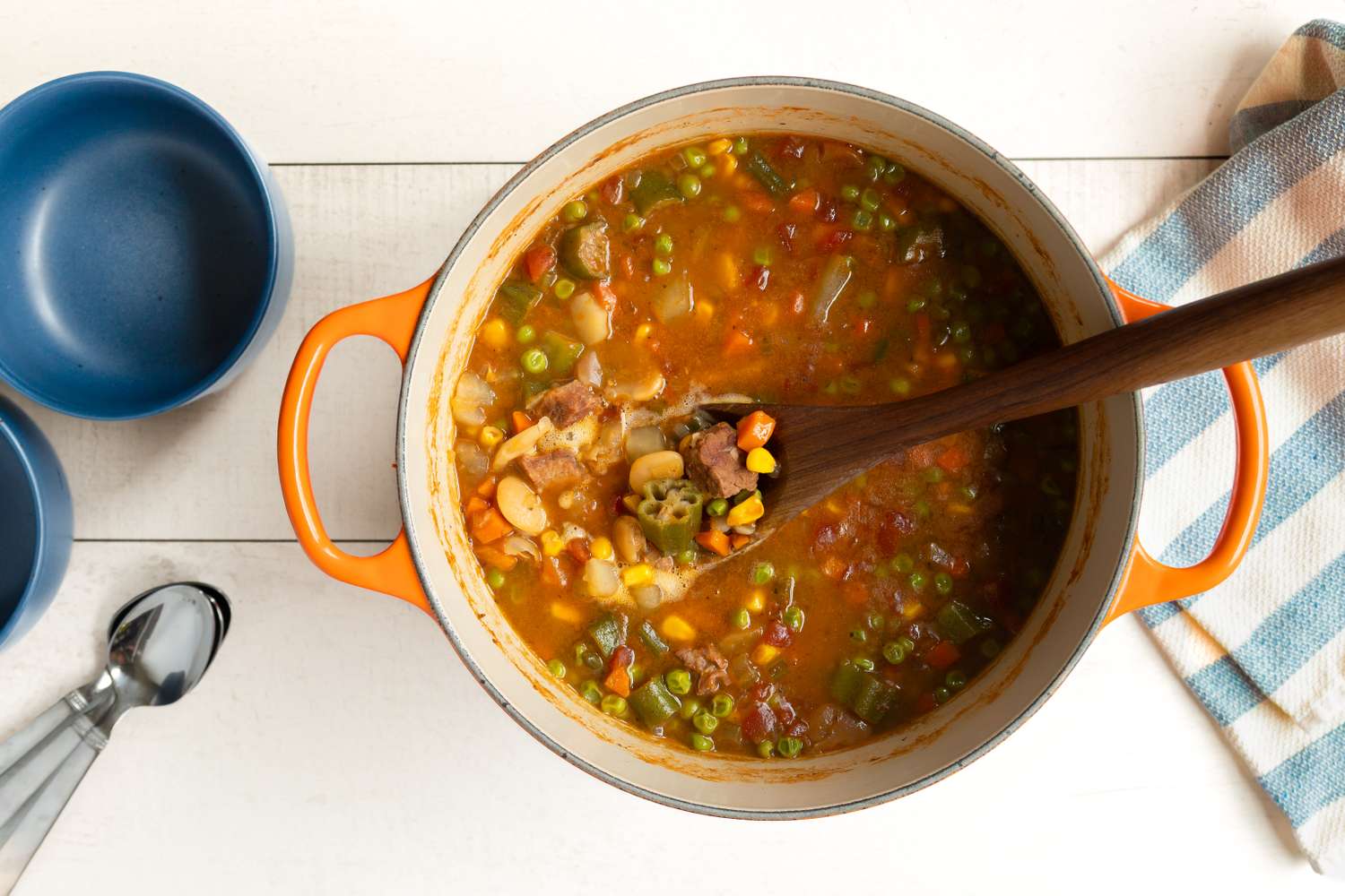 A pot of vegetable soup with a ladle next to empty bowls and spoons